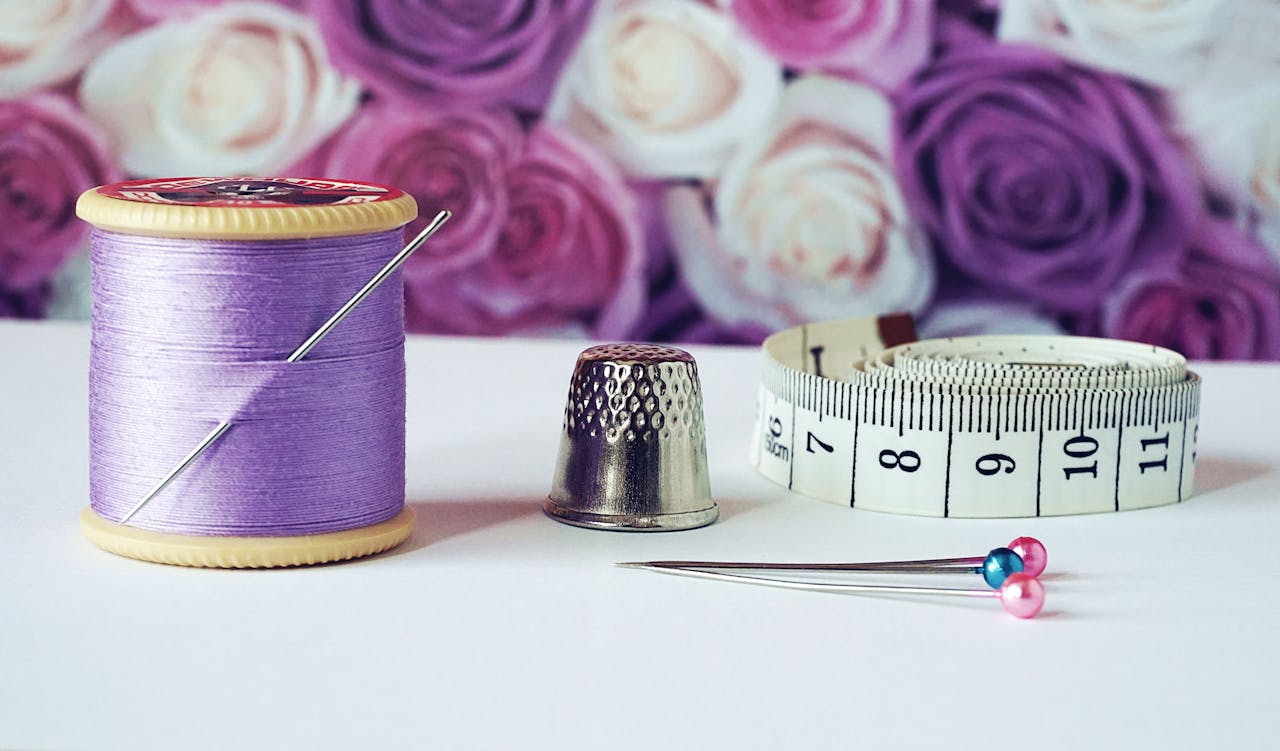 services-04 A close-up of sewing tools including lilac thread, a thimble, tape measure, and colorful pins with a rose backdrop.