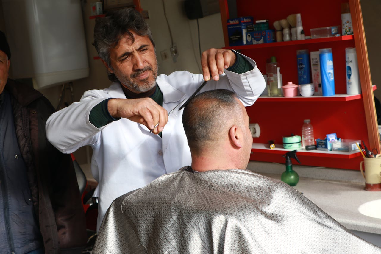 services-02 A barber skillfully cuts a customer's hair in a cozy, well-stocked shop.